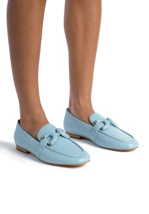 Mocassini in pelle azzurro chiaro con punta arrotondata, cinturino prominente e accessorio circolare in metallo. Suola piatta e texture liscia e lucida.
