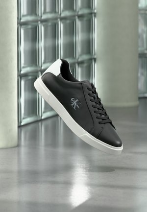 Baskets basses noires avec semelle blanche et languette de talon, logo Calvin Klein sur le côté, présentées en lévitation devant un mur en blocs de verre.