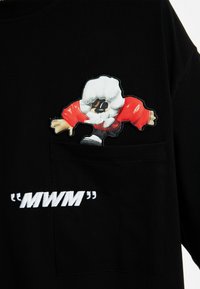 Camiseta negra con un personaje de dibujos animados en una chaqueta roja bordado en el bolsillo del pecho y el texto blanco "MWM" debajo.