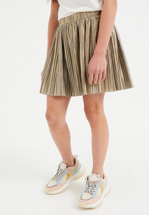 Fille portant une jupe plissée métallique dorée et des baskets épaisses multicolores, debout devant un fond uni.