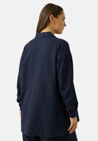 Granatowa lniana koszula o luźnym kroju, z kołnierzykiem w kształcie szpica, długimi rękawami i mankietami na guziki. Gładka tekstura, minimalistyczny design.
