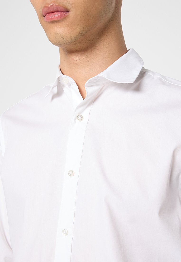 Close-up van een persoon in een wit overhemd met een open kraag tegen een effen achtergrond.