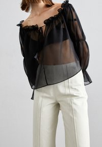 Nainen, joka pukeutuu läpinäkyvään mustaan off-shoulder-paitaan, jossa on röyhelöitä, ja valkoisiin korkeavyötäröisiin housuihin harmaata taustaa vasten.