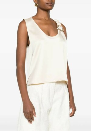 Top crema senza maniche con scollo rotondo e fibbia decorativa dorata sulla spalla sinistra, indossato con pantaloni bianchi larghi.