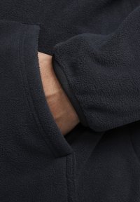 Fleecová bunda tmavě námořnické barvy s texturovaným povrchem. Má volný střih a ruku zasunutou do boční kapsy. Černé lemování na rukávu.