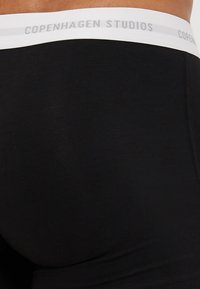 Schwarze Stoffshorts mit glatter Textur, ausgestattet mit einem weißen elastischen Bund, der mit grauer Schrift "COPENHAGEN STUDIOS" beschriftet ist.