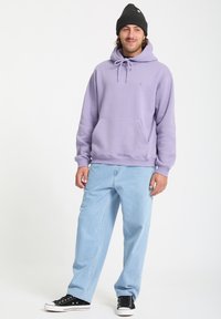Haut en coton violet avec une poche avant et un cordon de serrage ajustable. Associé à un pantalon en denim bleu clair et des baskets noires.