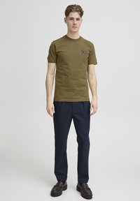Tricou scurt cu mânecă în verde măsliniu, cu guler rotund, având un mic logo negru pe pieptul stâng. Purtați-l cu pantaloni bleumarin și pantofi negri.