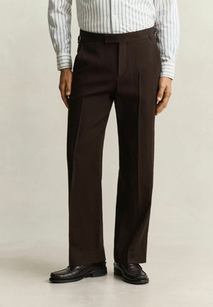 Man draagt donkerbruine, rechte broek gecombineerd met zwarte leren loafers en een wit overhemd met blauwe verticale strepen.