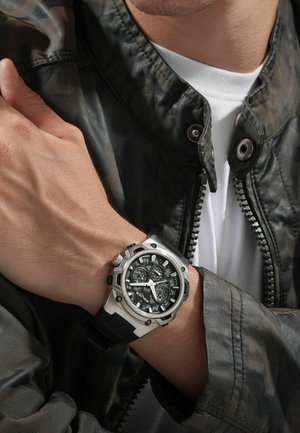 Black and silver watch with a textured dial, multiple sub-dials, and a black rubber strap, worn on a wrist with a camouflage jacket.