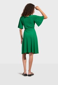 Femme aux cheveux bouclés portant une robe verte longueur genoux avec de larges manches et des sandales à plateforme noires, debout de dos avec le bras levé.