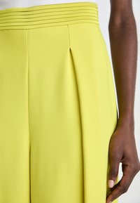 Geel op maat gemaakte broek met een geplooid ontwerp bij de taille, een gladde textuur en horizontale accenten langs de tailleband.