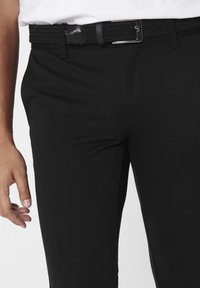 Zwarte pantalon met een gladde textuur, voorzien van een geweven zwarte riem en een zilveren metalen gesp. Close-up van het taillegebied en de hand.