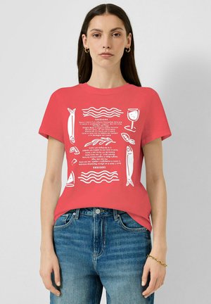 Kobieta ubrana w czerwoną koszulkę z białymi ilustracjami tematycznymi Sardynii: ryby, kieliszek wina, liście oraz tekst, w połączeniu z niebieskimi dżinsami i złotą biżuterią.