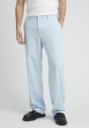 Homme portant un pantalon droit bleu clair, une chemise blanche rentrée, et des mocassins en cuir noir, debout avec une main dans la poche.