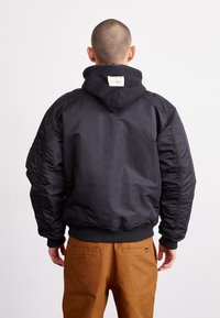 Black bomber jacket with a hoodie, featuring ribbed cuffs and waistband, sleek design, and a fabric tag on the back. Partnered with brown pants.