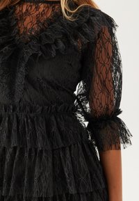 Svart spetsklänning med en figurnära bodice, volangkrage, genomskinliga långärmar och en lager på lager kjol med ett strukturerat spetspattern.