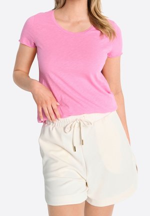 Femme portant un t-shirt rose vif à manches courtes et un short blanc à cordon, posant avec une main près de la taille.