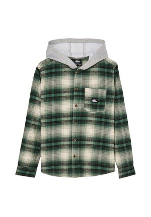Garçon souriant aux cheveux blonds bouclés, portant une chemise à carreaux verte, un jean bleu ample et des baskets noires, debout sur un fond clair et uni.
