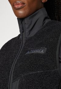 Black fleece vest with a high collar, textured material on front, smooth panels on sides, and a visible logo patch. Zip closure.
