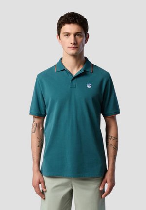 Uomo che indossa una polo turchese con rifiniture arancioni e pantaloni beige chiaro, in piedi contro uno sfondo grigio chiaro uniforme.