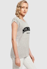 Grå kortermet T-skjorte med V-hals og svart "MUNDI MERCH" grafisk trykk, kombinert med hvite bukser. Myk stoffkvalitet, avslappet passform.