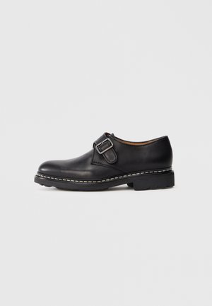 Sorte læder munkesko med sølvspænde og tyk sål med synlige hvide sømme på en ensfarvet baggrund.