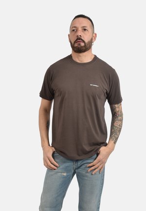 Uomo con barba e tatuaggi che indossa una maglietta marrone "Roy Roger's" e jeans azzurri, in piedi con le mani vicino alle tasche su sfondo bianco.