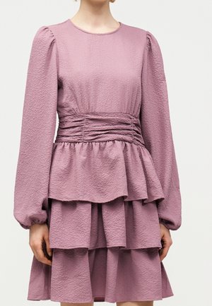 Kobieta w długim, teksturowanym, fioletowym (mauve) ubraniu z bufkami na rękawach, marszczoną talią i wielowarstwową spódnicą z falbanami.
