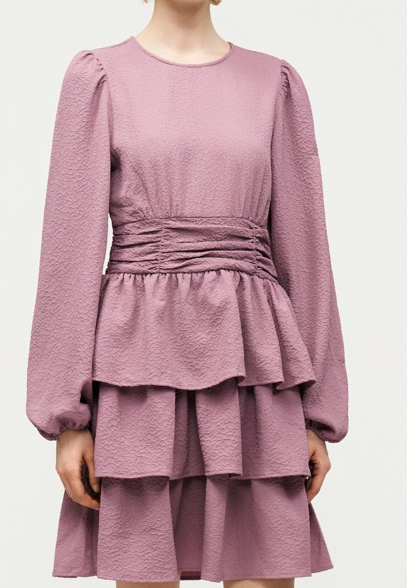 Femme portant une robe mauve texturée à manches longues, manches bouffantes, taille froncée et jupe à volants à étages.