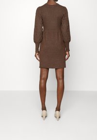 Abito in maglia marrone con maniche a sbuffo texturizzate, vita aderente e lunghezza fino a metà coscia. Abbinato a scarpe con tacco alte color beige e glitter.