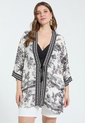 Kvinde iført kimono med sort-hvidt blomsterprint og geometrisk kant, sort top, hvide shorts og guldfarvede kvast-øreringe.