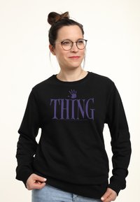 Žena s brýlemi a drdolem, oblečená v černé mikině s fialovým textem "THING" a otiskem ruky, stojí na jednobarevném pozadí.