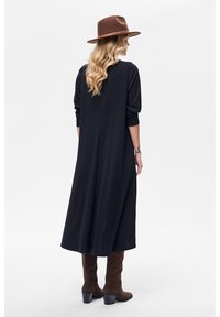 Długa czarna sukienka z długimi rękawami, gładka tkanina i luźny krój. Do tego brązowy kapelusz i brązowe zamszowe kozaki za kolano.