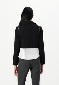 Kuttet svart jakke med hvit peplum-kant; har sølvfarget hardware på skuldrene; glatt tekstur og skreddersydd design.