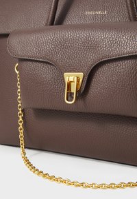 Sac à main en cuir texturé marron avec un fermoir triangulaire doré et une bandoulière en chaîne. Il possède une fermeture à rabat et un logo embossé sur le devant.