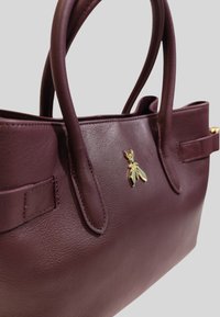 Borsa in pelle marrone scuro con doppio manico e un piccolo emblema d'ape dorata sul davanti, su uno sfondo neutro.