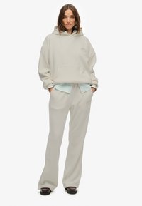 Felpa beige chiaro con tasca anteriore e logo ricamato, abbinata a pantaloni della tuta coordinati. Il materiale sembra morbido e confortevole.