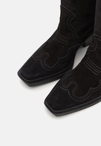 Black suede cowboy boots with a square toe, detailed stitching, and a scalloped design along the shaft. Embossed leather accents visible.
