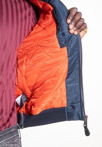 Main noire tenant ouverte une veste bleu marine montrant une doublure matelassée orange, personne portant une chemise rayée bordeaux et un jean gris.