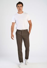 Bruine broek met een getailleerde pasvorm, achterzakken, en een zilveren knoop, gecombineerd met een effen wit T-shirt met ronde hals en witte sneakers.