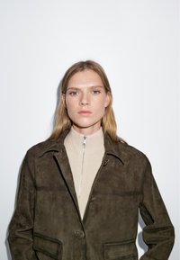 Giacca in suede marrone con colletto, pattina frontale e due tasche. Indossata sopra un dolcevita beige a coste. Design minimalista.