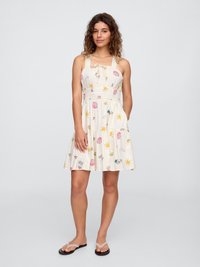 Jonge vrouw in een mouwloze witte zomerjurk met kleurrijke zon- en schelppatronen, draagt zwarte teenslippers en staat tegen een effen achtergrond.