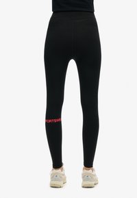 Leggings neri realizzati in tessuto elasticizzato, con vita alta e un logo rosso "SPORTSWEAR" sulla parte inferiore della gamba sinistra.