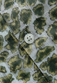 Tessuto leopardato verde e beige con una trama strutturata, caratterizzato da un bottone circolare a quattro fori cucito sulla superficie.