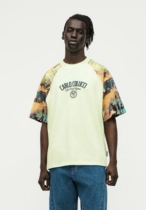 Junger Mann mit dunkler Haut und Dreadlocks, der ein hellgelbes T-Shirt mit mehrfarbigen Ärmeln und dem Schriftzug „Carlo Colucci“ trägt, kombiniert mit blauen Jeans.