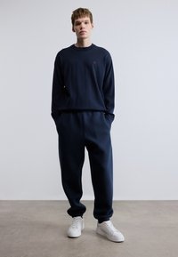 Σετ ναυτικού μπλε φούτερ και αντίστοιχων παντελονιών φόρμας από μαλακό ύφασμα, με μικρό λογότυπο στο στήθος. Ολοκληρώνεται με λευκά αθλητικά παπούτσια.