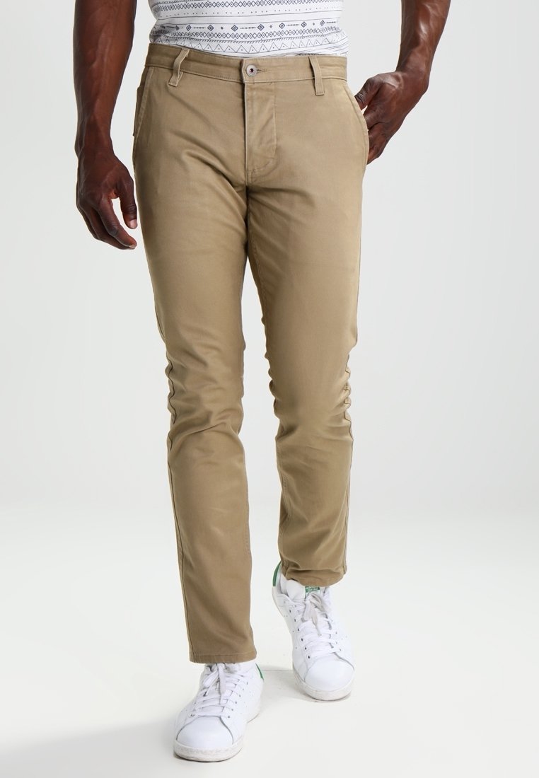 Homme portant un pantalon chino beige slim et des baskets blanches, marchant avec une main dans la poche sur un fond uni.