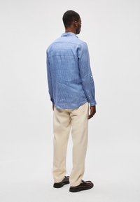 Ριγέ μπλε πουκάμισο από ελαφρύ ύφασμα, με γιακά και κουμπωτές μανσέτες, φορεμένο με μπεζ παντελόνι χαλαρής γραμμής και σκούρα παπούτσια.