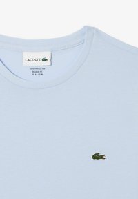 Camiseta de Pima algodón de color azul claro con cuello redondo, que presenta un pequeño logo de cocodrilo verde en el lado izquierdo del pecho. Diseño de ajuste regular.